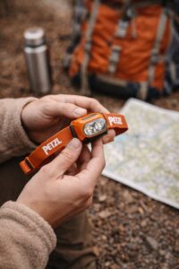 Retkeilijä pitelee otsalamppua käsissään, taustalla rinkka ja kartta retkeilyvarusteiden suunnittelua varten.