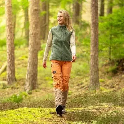 Nainen kävelee metsäpolulla vihreä liivi yllään ja vaaleanruskeat muunneltavat vaellushousut jalassa.